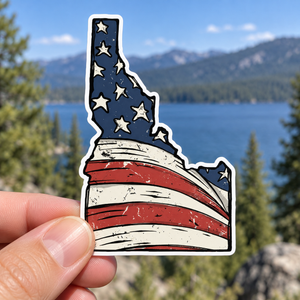 Idaho American Flag sticker held outdoors with a lake and evergreen trees in the background.